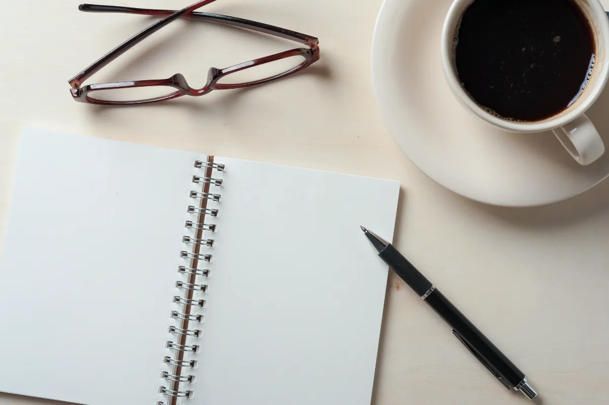a photo of a journal, a pen, a cup of coffee and eye glasses